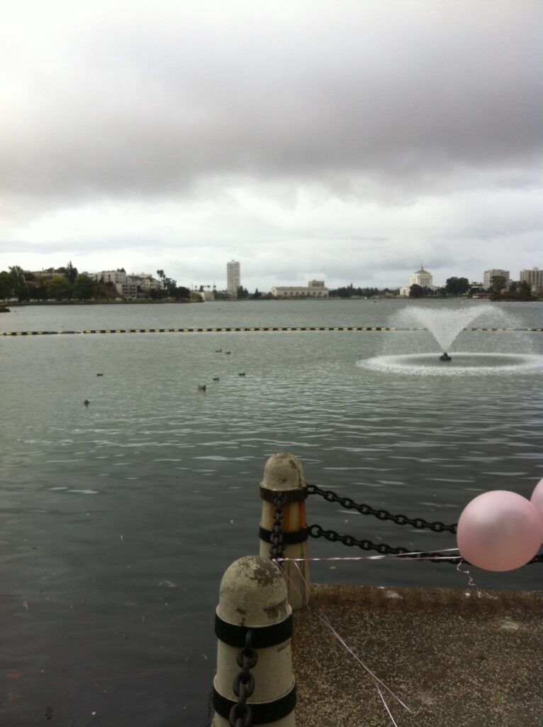 A portrait-orientation photograph of Lake Merritt in Oakland, California, as seen from the pergola. Photo taken in 2013. There is a fountain arcing water out over the lake in the middle ground, Oakland in the background, and a small platform ringed with balloons in the foreground.
