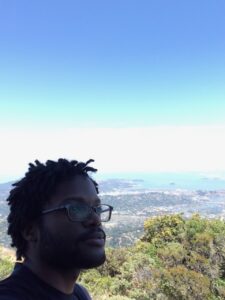 A photograph of David Brothers on top of a mountain. He's got short dreadlocks and glasses, and is looking up and to the right with nature behind him.