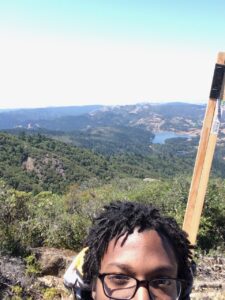 A photograph of David Brothers on top of a mountain. He's got short dreadlocks and glasses, and is carrying a bag of sticks on his back. Nature extends behind him.
