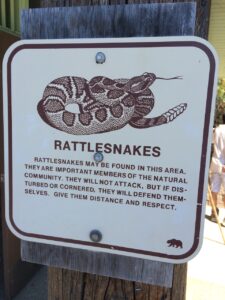 A photograph of a sign that warns hikers of rattlesnakes. The sign says, "Rattlesnakes may be found in this area. They are important members of the natural community. They will not attack, but if disturbed or cornered, they will defend themselves. Give them distance and respect."