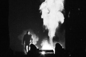 a black & white photograph of pro wrestler Tommaso Ciampa at AEW Dynasty 2026 in Vancouver, Canada. He's entering as flames and smoke billow behind him.